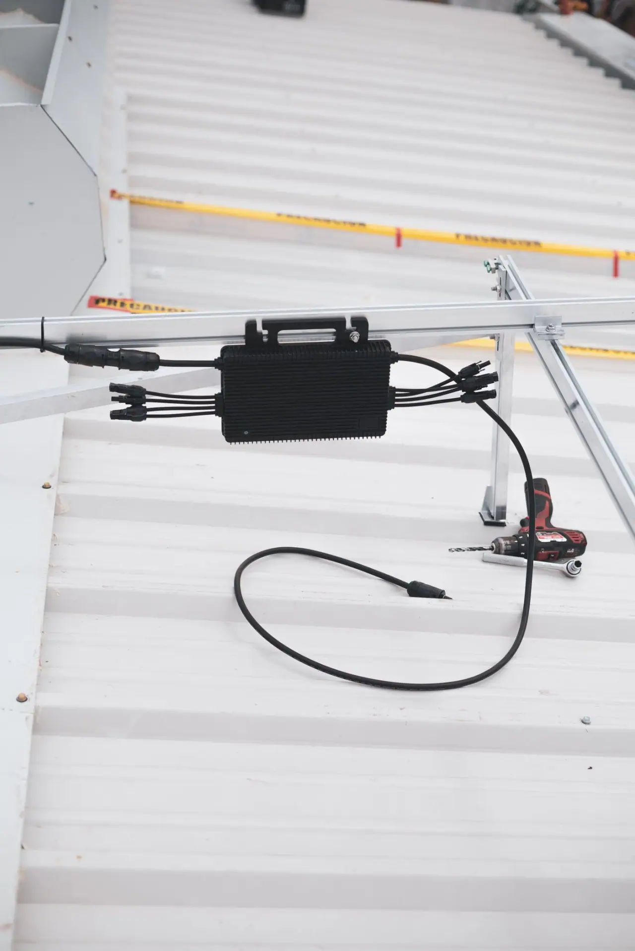 A module-level power electronics unit clamped to a mounting rail with its DC leads dressed along the purlin